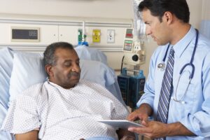A senior male patient in a hospital bed with a male doctor stood beside him holding a pad with a stethoscope around his neck. 