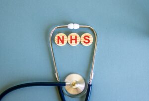 The letters 'NHS' on three separate wooden circular blocks in the middle of a stethoscope with a plain blue background. 